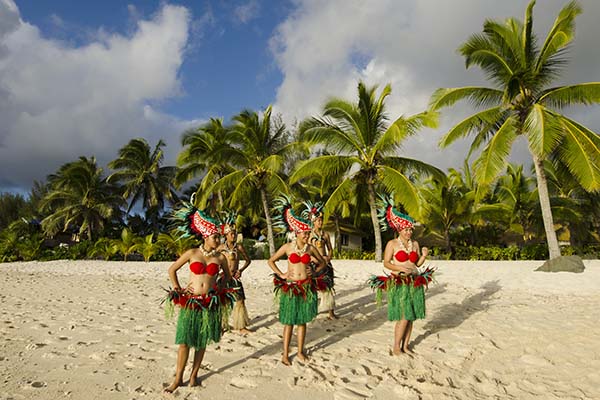 Group portrait of Polynesian Pacific Island Tahitian dance group in colorful costumes dancing on tropical beach with palm trees in the background.